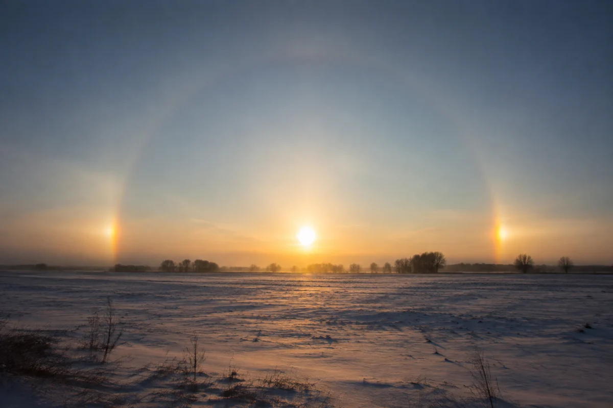 Illustration IA : Soleil bas accompagné de deux taches lumineuses dans un paysage enneigé.